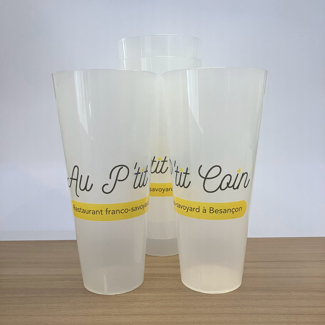 Trois gobelets réutilisables personnalisés pour le restaurant Au P'tit Coin, réalisés par l'agence Okami. Les gobelets arborent le logo et le nom du restaurant avec une bande jaune et le texte 'Restaurant franco-savoyard à Besançon'.